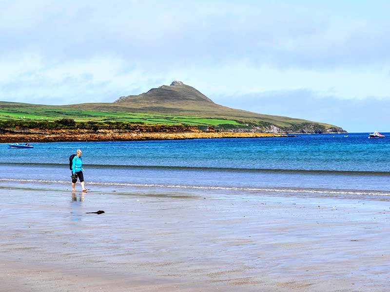 A woman walking in the water along a beach with a triangular green hill behind